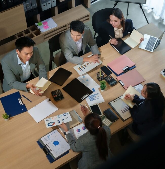 An overhead view of a virtual assistant team collaborating around a table with laptops, tablets, and data charts to coordinate remote client services.