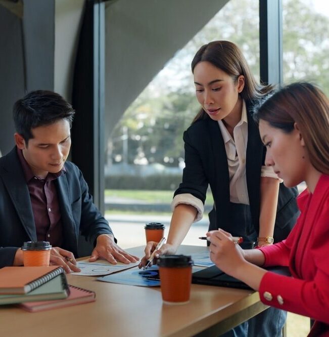 A team of three professional virtual assistants collaborating at a table, reviewing printed data charts and reports together in a modern office.