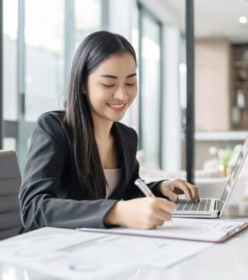 A professional female virtual legal assistant in a black blazer smiling while taking notes and working on a laptop in a modern office.