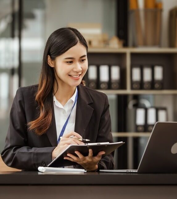 A professional female virtual legal assistant in a black blazer smiling while taking notes on a clipboard and looking at a laptop in an organized office.